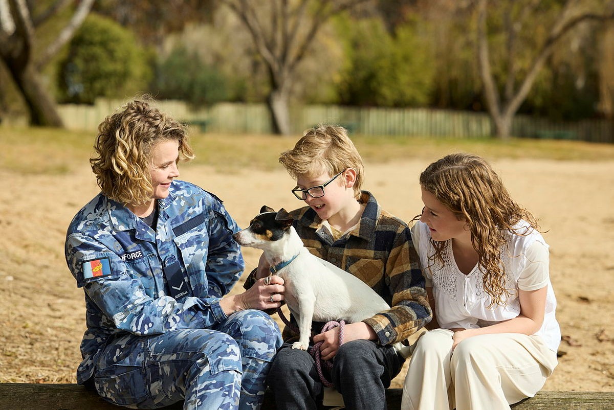 Photograph shows three people seated outdoors, gently holding and interacting with a small black-and-white dog. Left person is a woman, perhaps mother, and is wearing a blue camouflage uniform with patches, while centre and right are children wearing casual clothing in a warm, park-like setting.